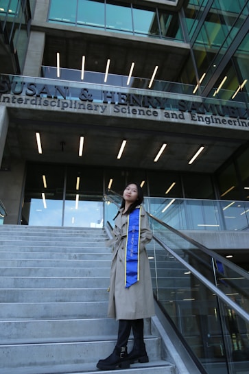 A person stands on a set of concrete stairs in front of a modern building with a clear glass facade. The building has a sign with the words 'Interdisciplinary Science and Engineering'. The individual is wearing a long beige coat and a blue sash that reads 'UC IRVINE'. They pose confidently, looking upwards.