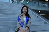 A young woman wearing a graduation gown sits on concrete steps. She has a blue graduation sash with yellow trim, displaying 'CLASS OF 2023'. The setting is outdoors with a modern building and glass railing in the background.