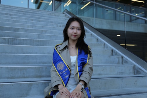 A young woman wearing a graduation gown sits on concrete steps. She has a blue graduation sash with yellow trim, displaying 'CLASS OF 2023'. The setting is outdoors with a modern building and glass railing in the background.