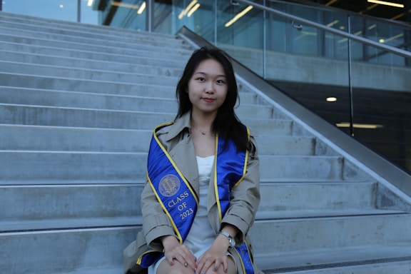 A young woman wearing a graduation gown sits on concrete steps. She has a blue graduation sash with yellow trim, displaying 'CLASS OF 2023'. The setting is outdoors with a modern building and glass railing in the background.
