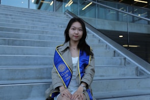 A young woman wearing a graduation gown sits on concrete steps. She has a blue graduation sash with yellow trim, displaying 'CLASS OF 2023'. The setting is outdoors with a modern building and glass railing in the background.