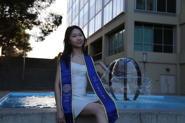 A young woman wearing a blue stole with 'UC Irvine' and 'Class of 2023' stands next to a water fountain. She is dressed in a white dress, and the setting appears to be outside a modern building with large windows.