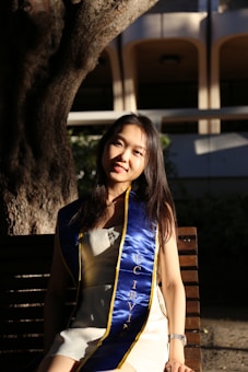 A person is sitting on a wooden bench under a tree, wearing a blue and yellow graduation sash that reads 'UC Irvine'. The background features architectural elements and is partially shaded.