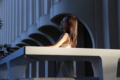A woman in a flowing white dress standing against a pale stone wall, bathed in natural light.