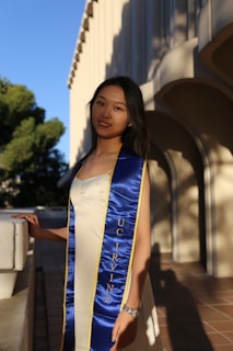 A woman stands outdoors wearing a blue graduation stole with yellow trim, embroidered with 'UC Irvine'. She is dressed in a light-colored dress, and there are architectural elements and trees in the background.