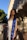 A woman stands outdoors wearing a blue graduation stole with yellow trim, embroidered with 'UC Irvine'. She is dressed in a light-colored dress, and there are architectural elements and trees in the background.
