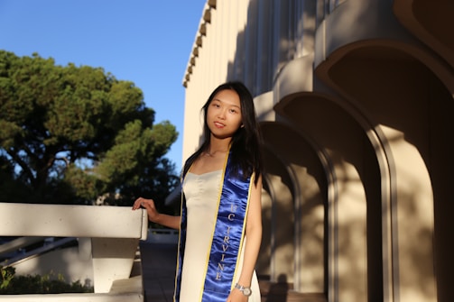 A young woman stands outdoors, wearing a blue and gold graduation stole over a light-colored dress. She is positioned next to a concrete railing, and behind her are large trees and a modern building with arches. The sunlight casts soft shadows on the building's walls.