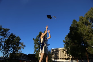 A person is in a graduation gown with a stole, joyfully throwing a graduation cap into the air. The background shows tall trees and a modern building, all under a clear blue sky.