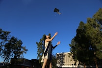A person in a graduation gown and cap is joyfully throwing their cap into the air. The setting is an outdoor area with tall trees and a clear, blue sky. In the background, there is a multi-storied building with distinctive architecture.