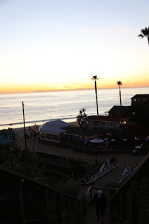A coastal scene at sunset with a calm ocean and sky painted in hues of orange and yellow. Palm trees dot the landscape while a beachfront area with buildings and outdoor seating is visible. People can be seen walking along the beach, and some lights are strung up, adding a warm glow to the structures.
