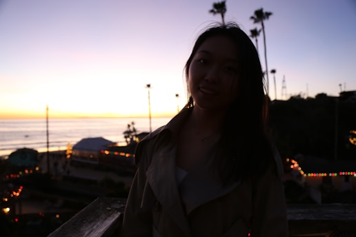 A silhouette of a person standing on a wooden structure during sunset. The background features the ocean and a colorful sky transitioning from yellow to deep blue. There are string lights illuminating parts of the surroundings, and palm trees are visible against the horizon.