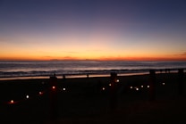 A serene beach at sunset with vibrant colors in the sky fading from blue to orange. Silhouettes of people can be seen along the shoreline, enjoying the peaceful atmosphere. String lights are softly glowing along a fence that runs parallel to the beach.