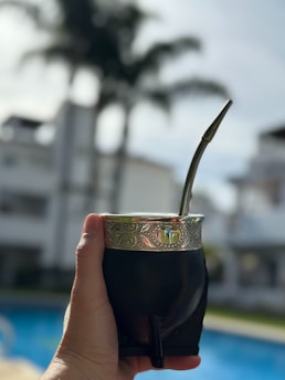 A hand holding a traditional mate cup with a metal straw, featuring ornate engravings on the rim. The background includes blurry residential buildings, palm trees, and a swimming pool, giving a sense of being outdoors on a sunny day.