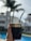 A hand holding a traditional mate cup with a metal straw, featuring ornate engravings on the rim. The background includes blurry residential buildings, palm trees, and a swimming pool, giving a sense of being outdoors on a sunny day.
