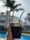 A hand holding a traditional mate cup with a metal straw, featuring ornate engravings on the rim. The background includes blurry residential buildings, palm trees, and a swimming pool, giving a sense of being outdoors on a sunny day.