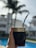 A hand holding a traditional mate cup with a metal straw, featuring ornate engravings on the rim. The background includes blurry residential buildings, palm trees, and a swimming pool, giving a sense of being outdoors on a sunny day.
