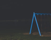 A dimly lit playground featuring a blue metal swing set with multiple swings. The surrounding area appears to be a grassy field with a dark, overcast atmosphere.
