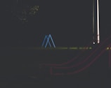 A dimly lit playground scene features a metal swing set in blue at the center. The environment suggests it is nighttime, with minimal lighting and shadows. A red slide is partially visible on the right side, while the surrounding area is largely enveloped in darkness.