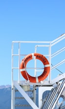 A bright orange life preserver is mounted on the white railing of a boat deck. The background features a clear blue sky with mountains visible in the distance. The life preserver is marked with the text 'Victoria Andina 1175'.