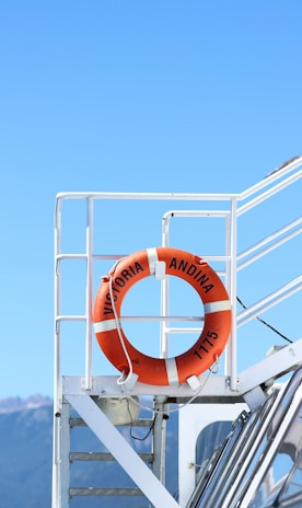 A bright orange life preserver is mounted on the white railing of a boat deck. The background features a clear blue sky with mountains visible in the distance. The life preserver is marked with the text 'Victoria Andina 1175'.