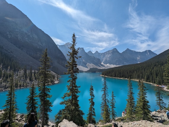 A picturesque landscape featuring a stunning turquoise lake surrounded by tall, evergreen trees and towering mountain peaks. The sky is clear with wispy clouds, adding to the serene and majestic atmosphere of the scene.