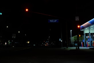 A nighttime street scene dominated by darkness with sporadic streetlights and traffic signals. A red traffic light commands attention in the upper center, while a green arrow traffic signal is visible to the left. On the right, a lit-up gas station displays a canopy with bright blue and white lights, and gas pumps are visible beneath it. Signs such as '166th St' and 'Truck Route' are readable, suggesting a minor intersection.