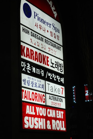A vertical sign displaying multiple business names in both English and Korean. The businesses include a spa, restaurant, karaoke bar, church, tailoring and alteration services, and a sushi restaurant. The sign is brightly lit with a mix of blue, red, and white colors.