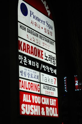 A vertical sign displaying multiple business names in both English and Korean. The businesses include a spa, restaurant, karaoke bar, church, tailoring and alteration services, and a sushi restaurant. The sign is brightly lit with a mix of blue, red, and white colors.