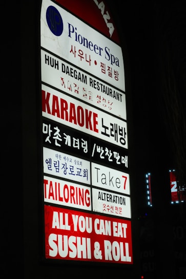 A vertical sign displaying multiple business names in both English and Korean. The businesses include a spa, restaurant, karaoke bar, church, tailoring and alteration services, and a sushi restaurant. The sign is brightly lit with a mix of blue, red, and white colors.