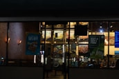A nighttime view of a restaurant exterior with promotional posters on the windows offering deals such as 'Buy One Get One 50% Off' and 'Buy 4 Get 1 Free'. Interior lighting reveals menu boards displaying various food items.