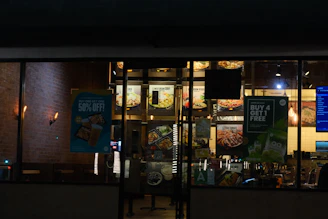 A nighttime view of a restaurant exterior with promotional posters on the windows offering deals such as 'Buy One Get One 50% Off' and 'Buy 4 Get 1 Free'. Interior lighting reveals menu boards displaying various food items.
