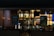 A nighttime view of a restaurant exterior with promotional posters on the windows offering deals such as 'Buy One Get One 50% Off' and 'Buy 4 Get 1 Free'. Interior lighting reveals menu boards displaying various food items.