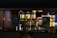 A nighttime view of a restaurant exterior with promotional posters on the windows offering deals such as 'Buy One Get One 50% Off' and 'Buy 4 Get 1 Free'. Interior lighting reveals menu boards displaying various food items.