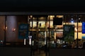 A nighttime view of a restaurant exterior with promotional posters on the windows offering deals such as 'Buy One Get One 50% Off' and 'Buy 4 Get 1 Free'. Interior lighting reveals menu boards displaying various food items.