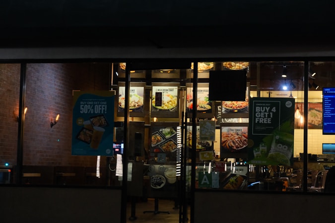 A nighttime view of a restaurant exterior with promotional posters on the windows offering deals such as 'Buy One Get One 50% Off' and 'Buy 4 Get 1 Free'. Interior lighting reveals menu boards displaying various food items.