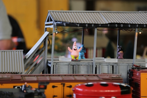 A detailed miniature scene featuring a colorful toy version of a cartoon character, standing on a platform with a corrugated metal roof. The character is pink with a distinctive star shape and is accompanied by a blue toy character in the background. Below the platform, a train model with orange and red hues is visible, along with a bright red object with black spots resembling a ladybug. The scene has a playful and whimsical feel.