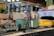 A model train set featuring a yellow train engine with 'Union Pacific' written on the side is on a track. In the foreground, a model military tank is visible on a flatbed car. The train is passing under a metal bridge structure which appears to be part of a larger model landscape, including buildings with sloped roofs and a tunnel entrance.