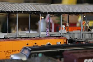 A detailed miniature model train scene featuring a tractor-trailer with a Union Pacific logo. Around it are small figurines of workers in various poses on a platform with a corrugated roof. A small purple creature stands on the orange boxcar, adding a quirky element to the scene.