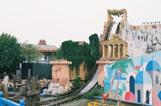 A theme park featuring a structure resembling ancient ruins next to a vibrant mural depicting Mediterranean architecture. The slide adorned with ivy descends from a height, suggesting a water ride. Bright colors and lush greenery surround the scene.