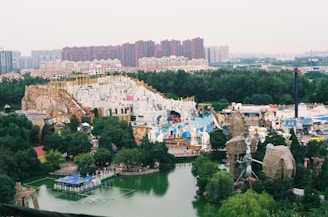 An aerial view of an amusement park with various structures and themed buildings surrounded by lush green trees. In the foreground, there is a small body of water with a bridge crossing over it. In the background, a dense line of modern high-rise buildings can be seen, portraying a cityscape.