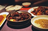 A table is set with a variety of Asian dishes, including a bowl of rich, dark glazed meat in a brown sauce, a plate of noodles with a golden sauce, and a dish of dumplings. The setting has a warm, ambient lighting that enhances the deep, rich colors of the food.