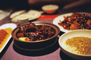 A table is set with a variety of Asian dishes, including a bowl of rich, dark glazed meat in a brown sauce, a plate of noodles with a golden sauce, and a dish of dumplings. The setting has a warm, ambient lighting that enhances the deep, rich colors of the food.