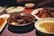 A table is set with a variety of Asian dishes, including a bowl of rich, dark glazed meat in a brown sauce, a plate of noodles with a golden sauce, and a dish of dumplings. The setting has a warm, ambient lighting that enhances the deep, rich colors of the food.