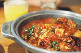 A close-up view of a savory dish featuring a rich tomato sauce with chunks of vegetables and herbs. The dish is served in a rustic, round pot with a wooden spoon, suggesting a homely or traditional meal. A glass of orange juice is visible in the background on a wooden table.