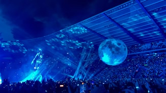 A vibrant concert scene with a large crowd under a stadium roof. A holographic figure is illuminated on stage, accompanied by a giant spherical moon structure. Bright blue lighting and laser beams create a dynamic and futuristic atmosphere.