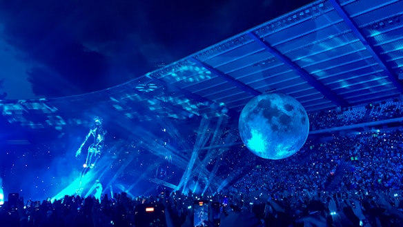 A vibrant concert scene with a large crowd under a stadium roof. A holographic figure is illuminated on stage, accompanied by a giant spherical moon structure. Bright blue lighting and laser beams create a dynamic and futuristic atmosphere.