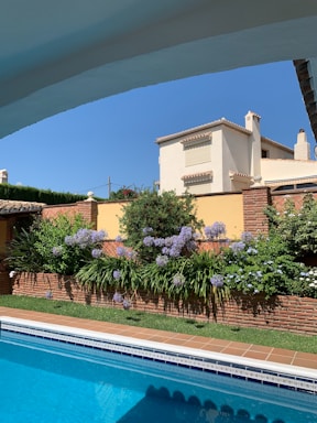 A picturesque scene of a luxurious house with a terracotta roof set against a clear blue sky. In the foreground, a swimming pool with clear blue water reflects the sky above. Surrounding the pool is a well-maintained garden with vibrant purple flowers and lush green foliage encased by a brick wall.
