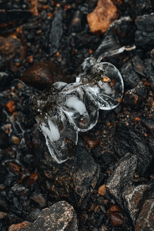A crystal-clear piece of ice rests on a bed of dark, jagged rocks and gravel. The ice is partially melted, with intricate patterns and smooth surfaces that catch the light. The stones vary in size and color, featuring shades of brown, black, and grey, creating a stark contrast with the ice.