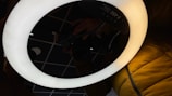 A circular LED ring light is illuminated, casting a soft glow. A reflection on a glossy surface shows a person's hand holding a camera. The background includes a grid pattern ceiling and part of a person wearing a yellow jacket.
