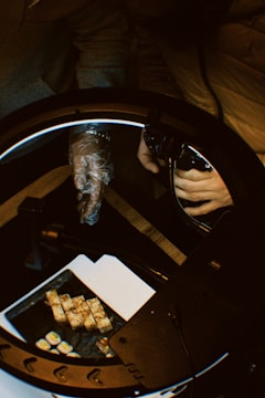 Two people are engaged in an activity involving a camera and a ring light, with one person wearing a glove and holding the light while the other manages camera settings. On the table, there is a tray containing pieces of sushi and some tofu-like food items, placed on a white surface.